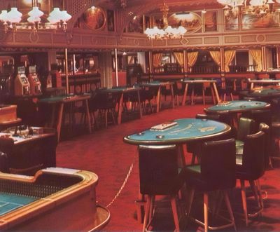 A restored 1954 brochure photograph of the Showboat casino floor with craps and blackjack tables, roulette, slot machines, and a gilded balcony rail