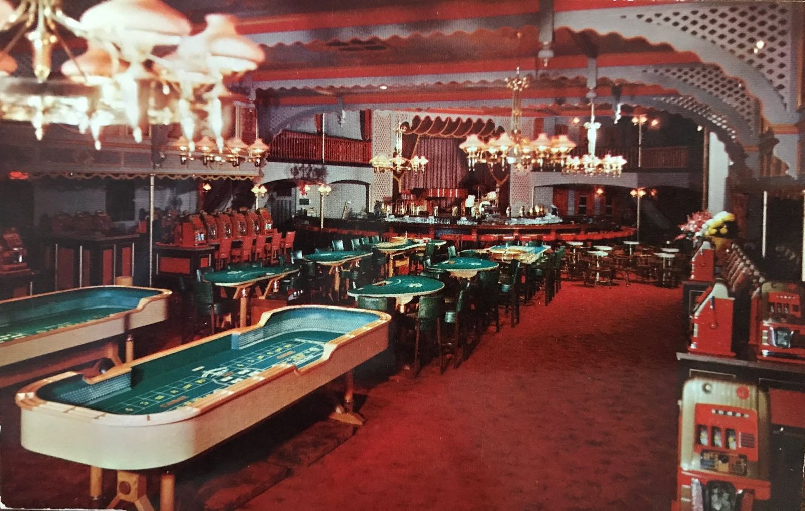 A 1950s color view of the Showboat casino floor: two craps tables and blackjack tables on red carpet, gas-lamp style chandeliers, scalloped balcony trim, slot machines along the right wall, and the curved bar in the distance