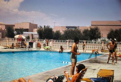 The Showboat pool in 1968 surrounded by webbed loungers and sunbathers, with a pink decorative breeze-block fence, a red and white striped umbrella, and salmon-pink buildings beyond