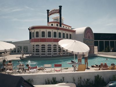 A restored 1950s photograph of the Showboat pool with sunbathers on director's chairs and loungers, two guests under a large white parasol, the white riverboat facade with red trim and twin black smokestacks behind, the SHOWBOAT bulb sign above, the ornate sunburst-arch SHOWBOAT entrance at right, and Bingo neon script on the dark casino block
