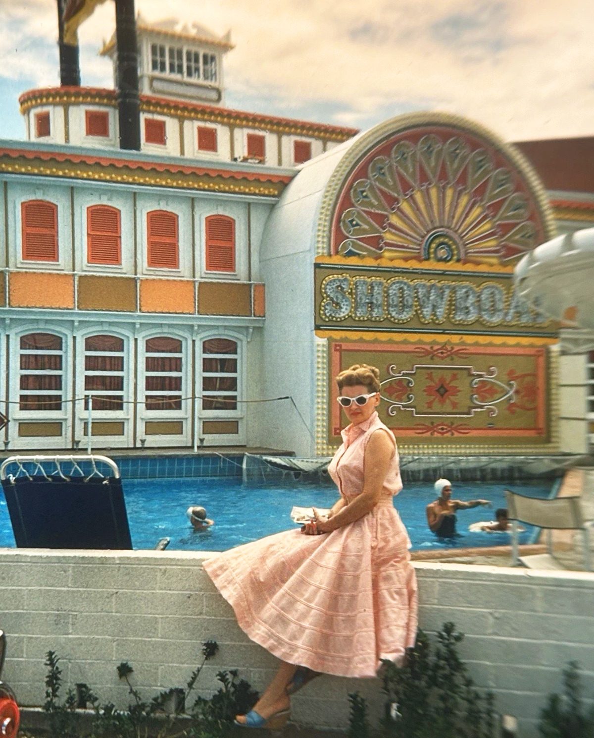 A woman in a pink 1950s circle dress and white cat-eye sunglasses sitting on the Showboat pool wall, with the ornate riverboat facade, coral sunburst arch, and chrome SHOWBOAT bulb letters behind her