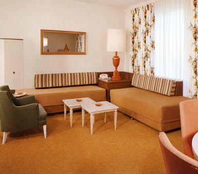 A restored 1950s Showboat brochure photograph of a suite living area with two ochre daybed sofas with striped bolsters, white marble nesting tables, a green armchair, floral drapes, and an orange table lamp beside a rotary telephone