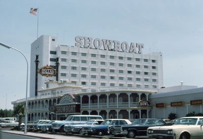 The white Showboat hotel tower in 1975 with SHOWBOAT letters across the roofline, an American flag above, the Hotel Showboat sign on a black smokestack, a gingerbread veranda with Showboat Bowl, Keno, Bowling, and Casino signs, and 1970s cars parked in front